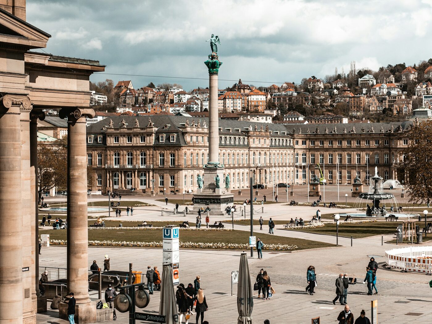 Stuttgarter Schlossplatz mit Jubiläumssäule, der Barockfassade des Neuen Schlosses und den Häusern der Halbhöhenlagen unter bewölktem Himmel